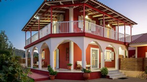 A two-story building with a red and white color scheme, featuring a balcony on the upper floor, arched doorways, and potted plants on the ground floor. - Diogenis House (Zakynthos)