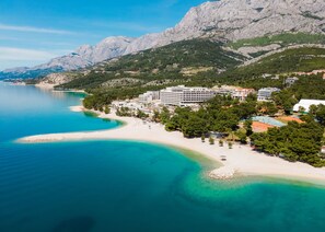 A coastal hotel with a beach, mountains, and clear blue waters.