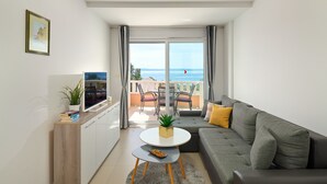 A modern living room with a grey sofa, a white coffee table, and a TV on a wooden cabinet. A balcony with a view of the sea is visible through the glass doors. - Sun Spalato Villa Ivano (Makarska)