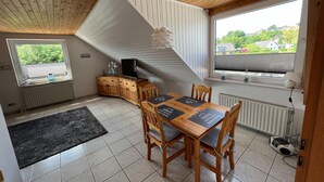 Un coin repas avec une table et des chaises en bois, un téléviseur posé sur un meuble, et une vue sur la verdure à l’extérieur.