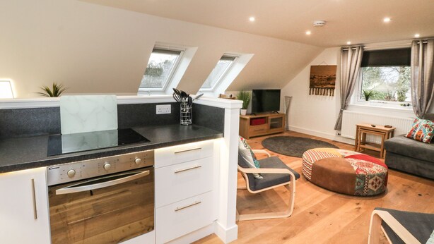 A modern kitchen with a black countertop, white cabinets, and a built-in oven. There is a dining area with a wooden table and chairs, and a cozy seating area with a patterned ottoman. - Taorbeag at Taormina (Fort William)
