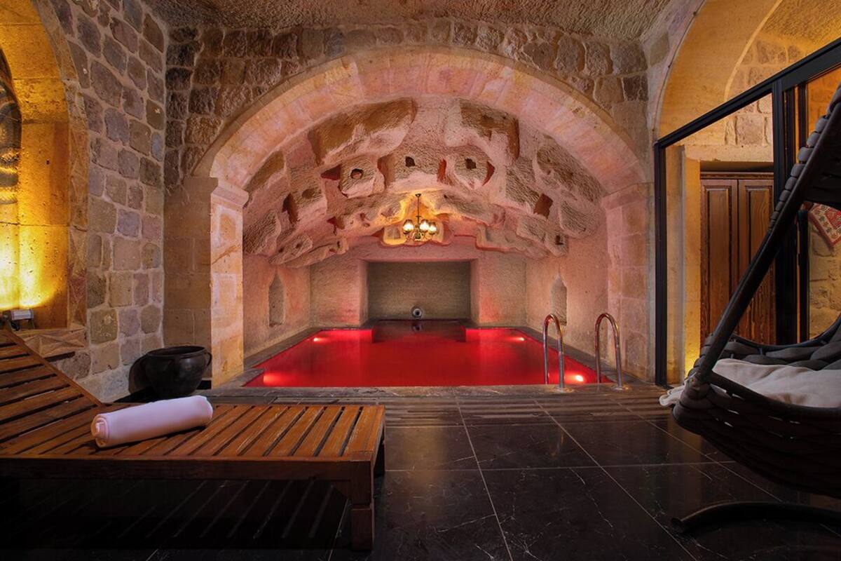 Ein Zimmer mit Steinwänden, einer rot beleuchteten Whirlwanne, Holzbanken und einem Hängesessel.