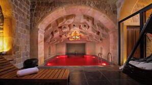 A stone-walled room with a red-lit hot tub, wooden benches, and a hanging chair.