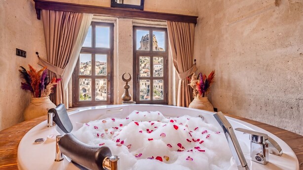A room with a bathtub filled with water and rose petals, a window with a view of buildings, and decorative vases.