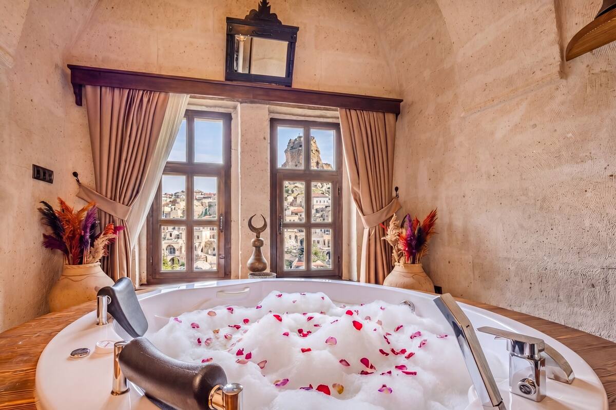 Ein Zimmer mit einer mit Wasser und Rosenblättern gefüllten Badewanne, einem Fenster mit Blick auf Gebäude und dekorativen Vasen.
