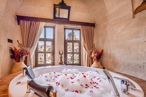 Una habitación con una bañera llena de agua y pétalos de rosa, una ventana con vistas a edificios y jarrones decorativos.