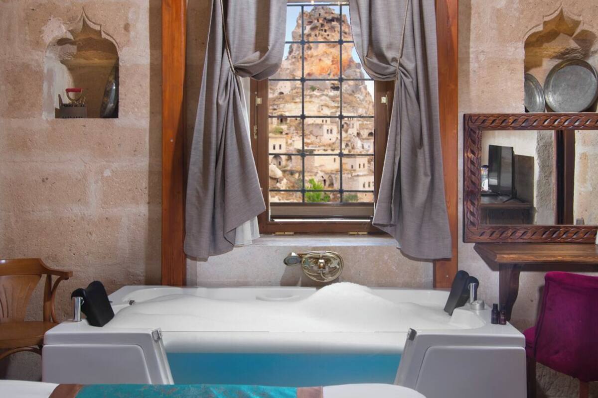 Ein Badezimmer mit einer Badewanne, einem Fenster mit Vorhängen, einem Holzstuhl, einem Spiegel und Blick auf eine felsige Landschaft.
