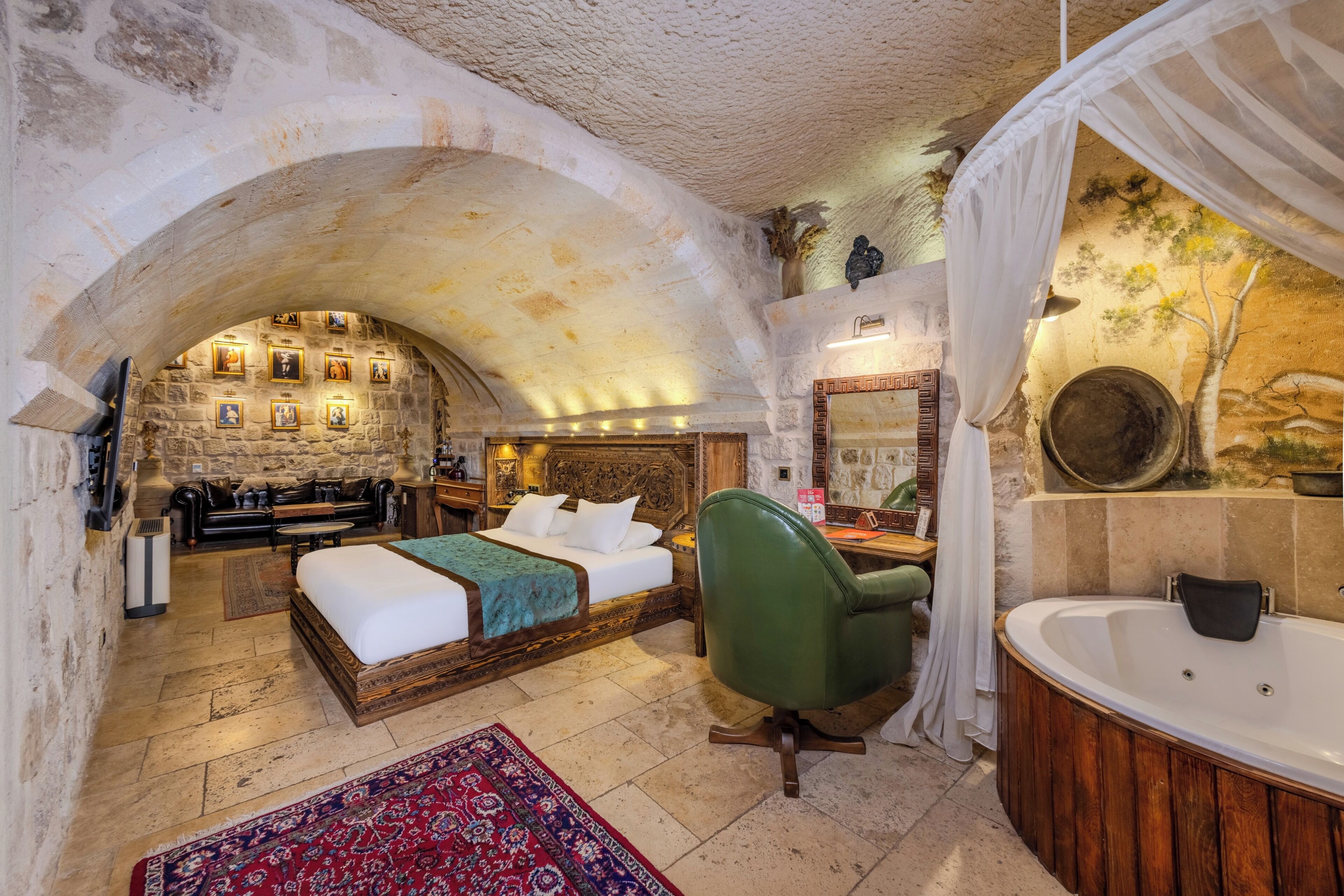 A bedroom with a stone archway, a bed, a green chair, a large bathtub, and a stone wall with framed pictures.