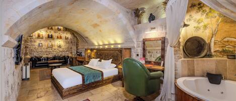 A bedroom with a stone archway, a bed, a green chair, a large bathtub, and a stone wall with framed pictures.
