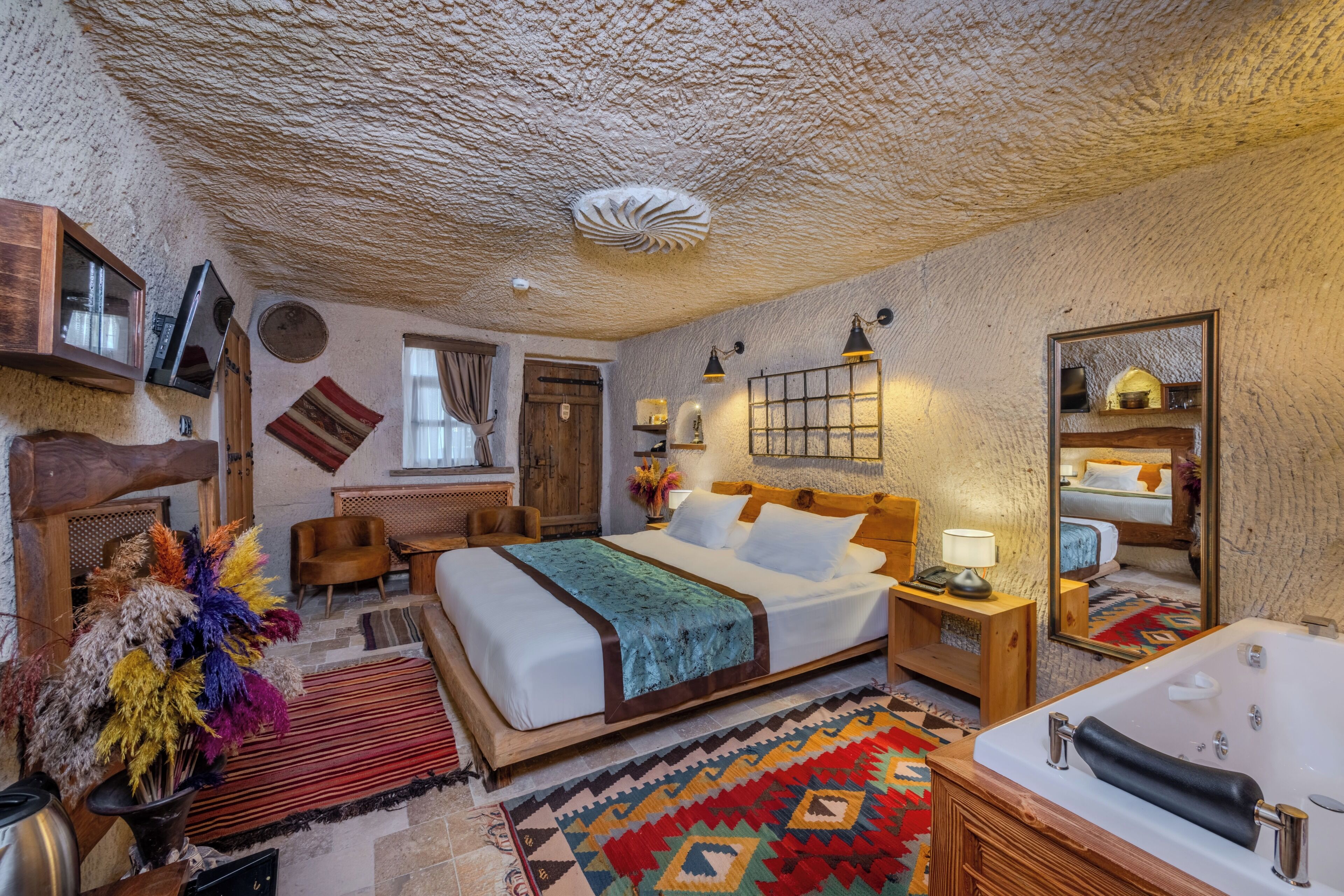 A room with a large bed, a wooden headboard, a nightstand with a lamp, a mirror, a bathroom with a bathtub, and a decorative rug.