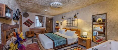 A room with a large bed, a wooden headboard, a nightstand with a lamp, a mirror, a bathroom with a bathtub, and a decorative rug.