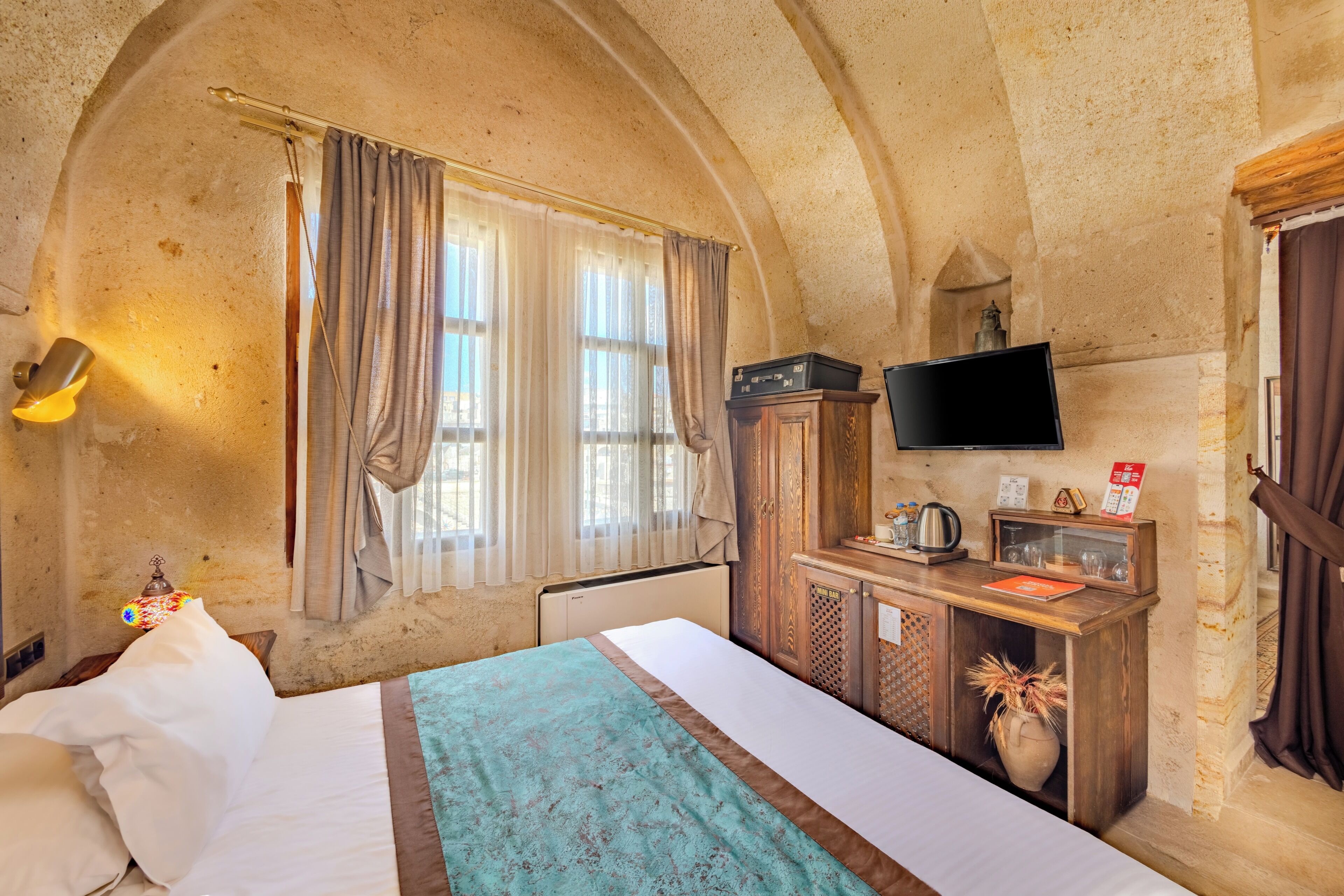 A room with a bed, a desk with a television, a radiator, and a window with curtains.