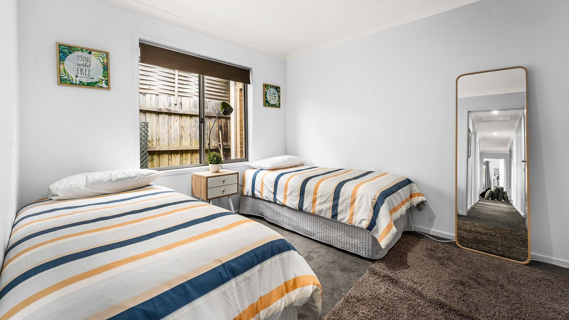 A bedroom with two beds, a window with a view of a wooden fence, a bedside table, and a mirror.
