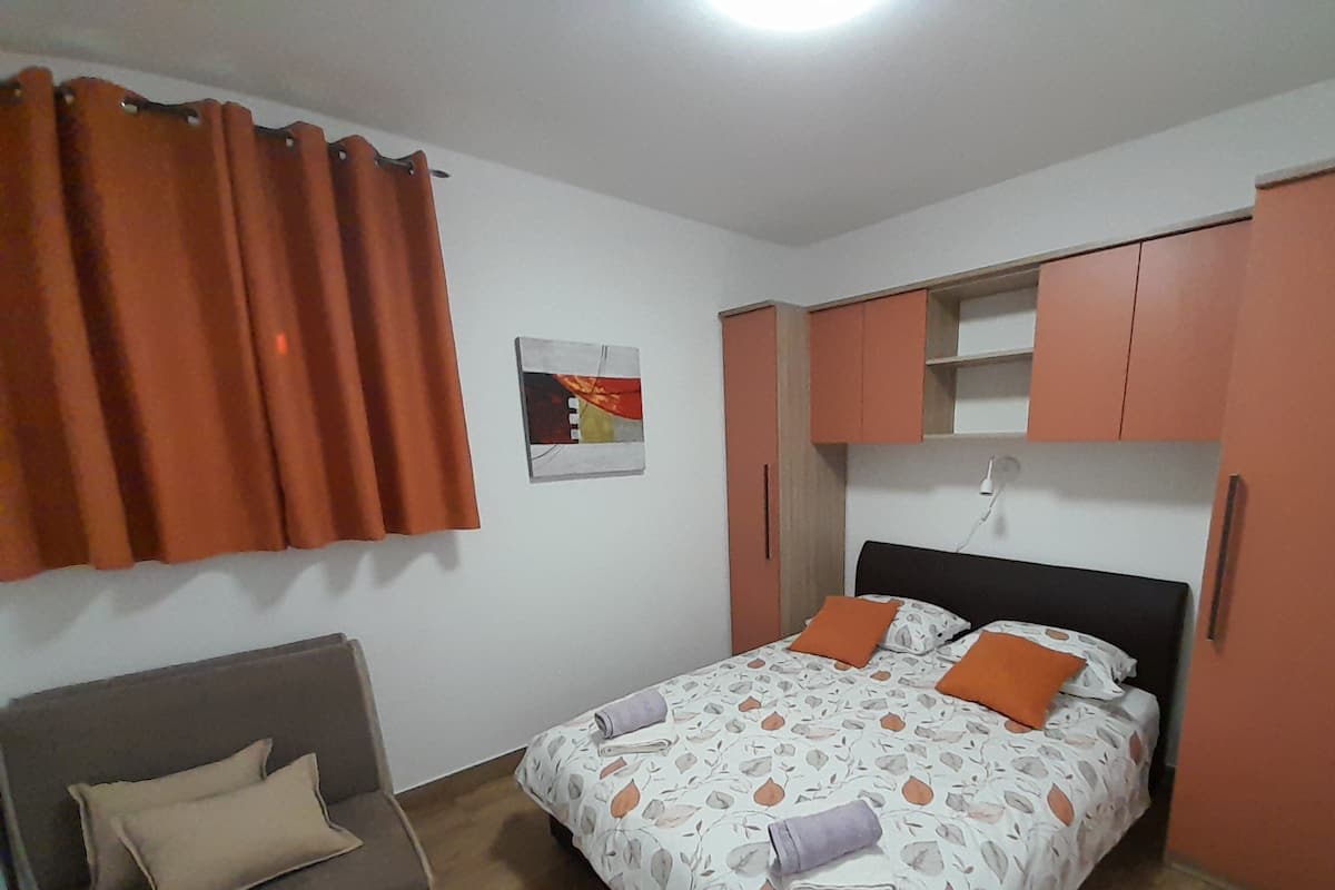 A bedroom with a bed, a grey sofa, orange curtains, and a wall-mounted shelf.