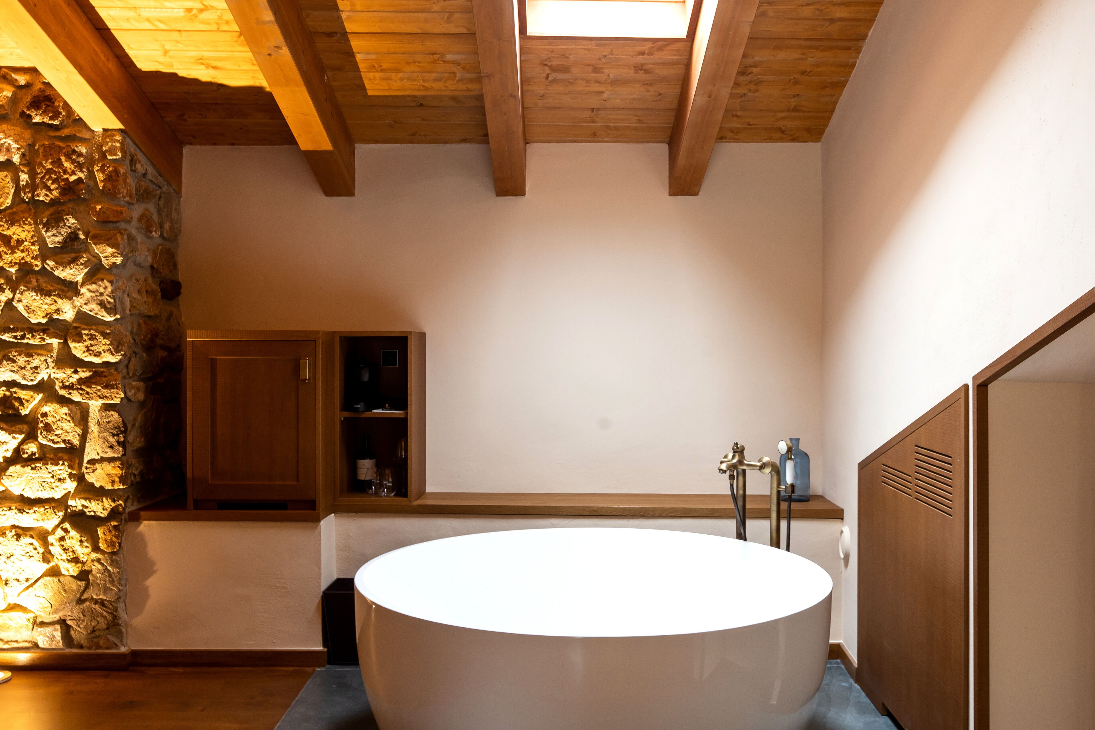 Un baño moderno con una bañera independiente, pared de piedra, vigas de madera y un estante empotrado con artículos de tocador.