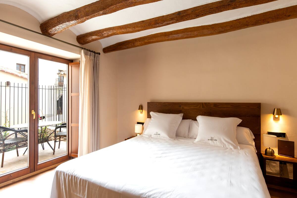 A bedroom with a wooden ceiling, a large bed with white bedding, two bedside lamps, and a view of an outdoor patio with a table and chairs.