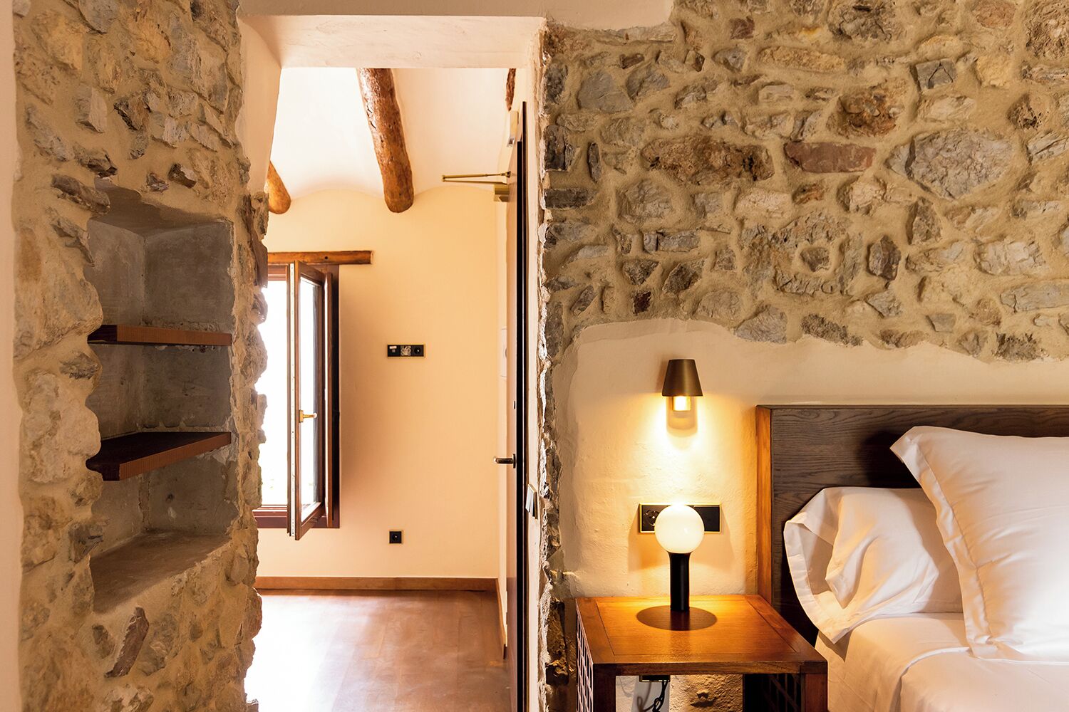 Un dormitorio con paredes de piedra, estructura de cama de madera, una mesita de noche con lámpara y ventana con vista al interior.