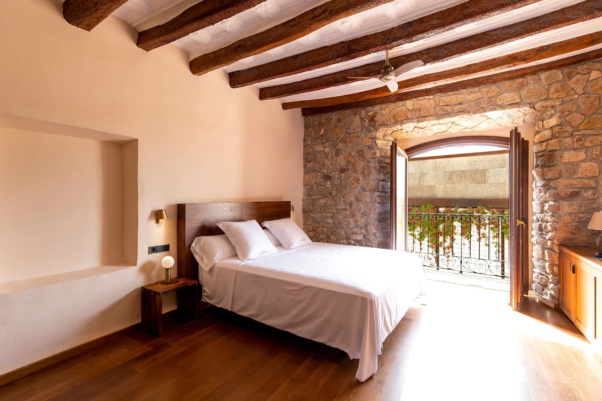 A bedroom with a stone wall, wooden beams, a bed, a nightstand, and a view of a balcony with plants.