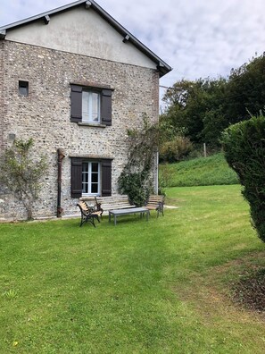 Une maison en pierre à deux étages, avec une cour verdoyante, un banc et une petite table.