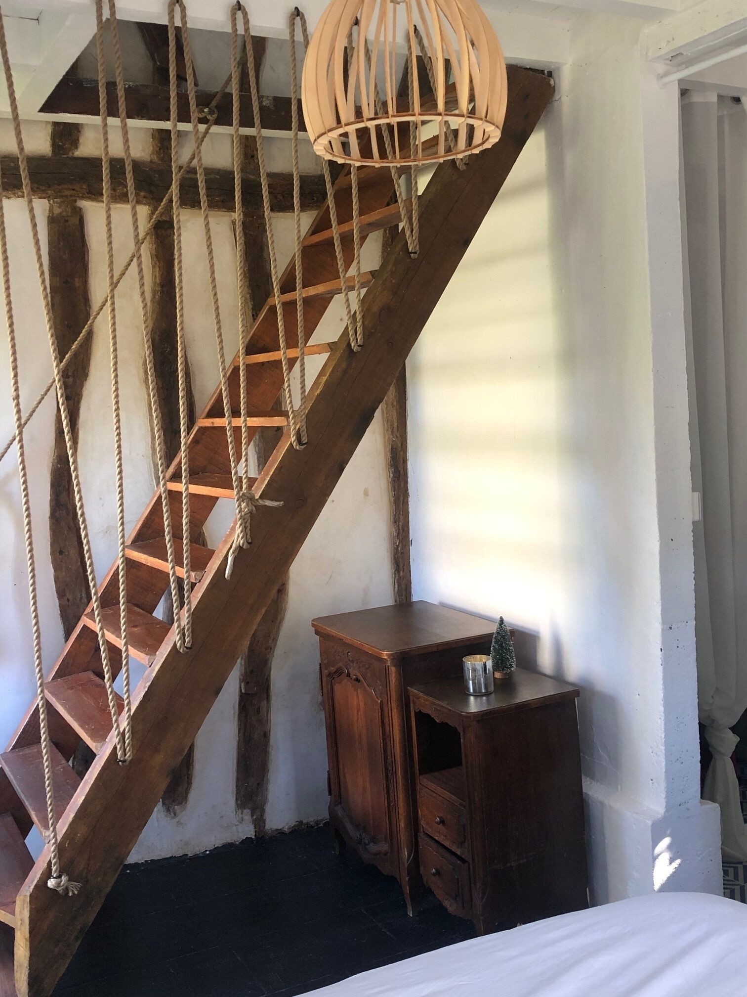 Un escalier en colimaçon en bois avec des rampes en corde, une petite armoire en bois et un luminaire suspendu.