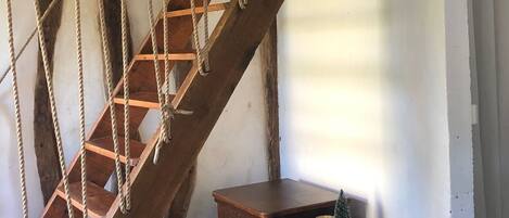 Un escalier en colimaçon en bois avec des rampes en corde, une petite armoire en bois et un luminaire suspendu.