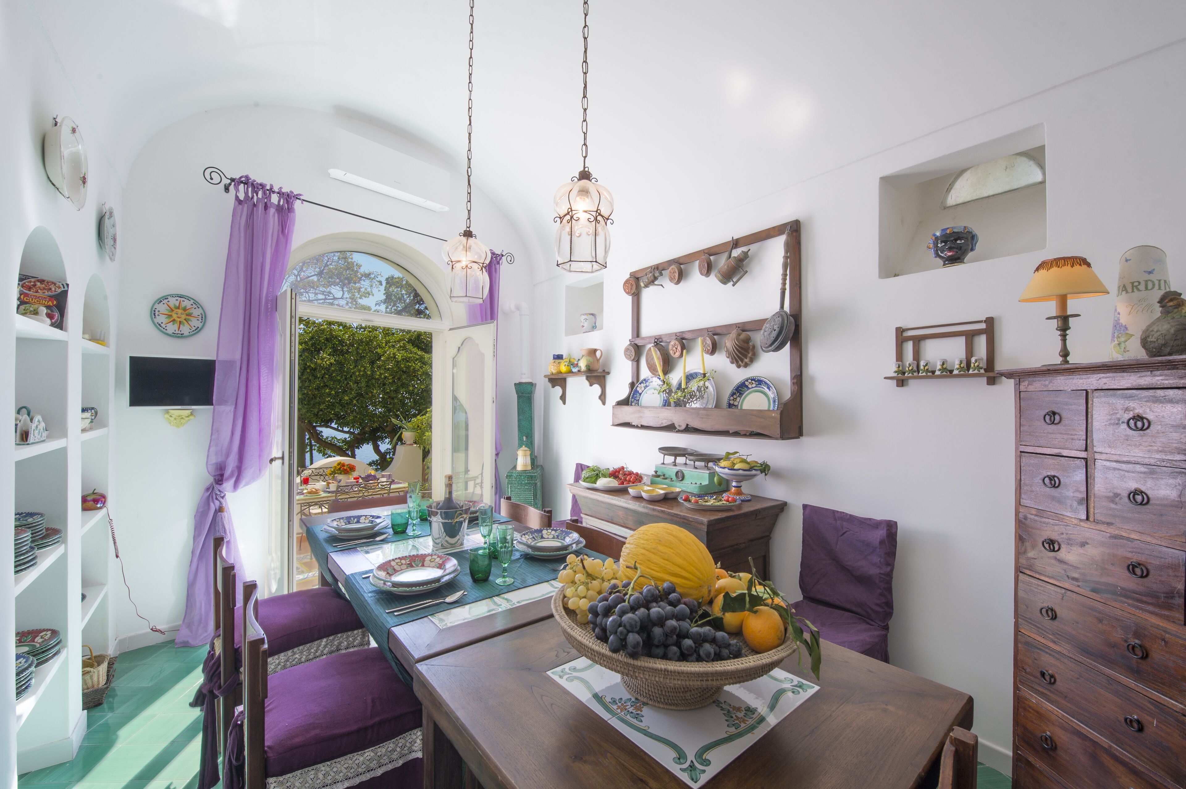 A dining area with a table set for two, featuring a fruit bowl, a wooden cabinet, and a window with purple curtains.