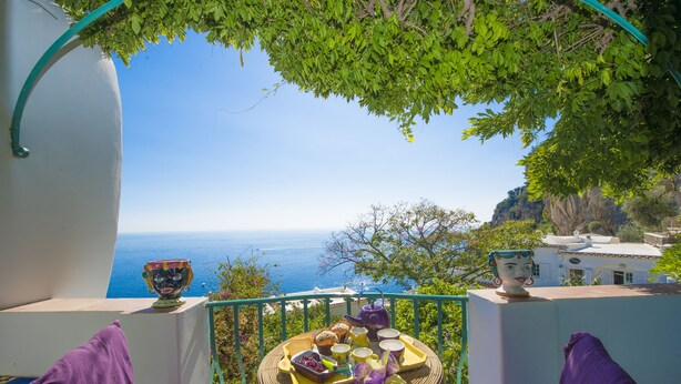 A balcony with a view of the sea, a table with food and drinks, and a decorative mask. - Casa del Fico Dimora storica Positano (Positano)