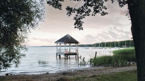 Un quiosco sobre pilares a orillas de un lago tranquilo con barcos al fondo.