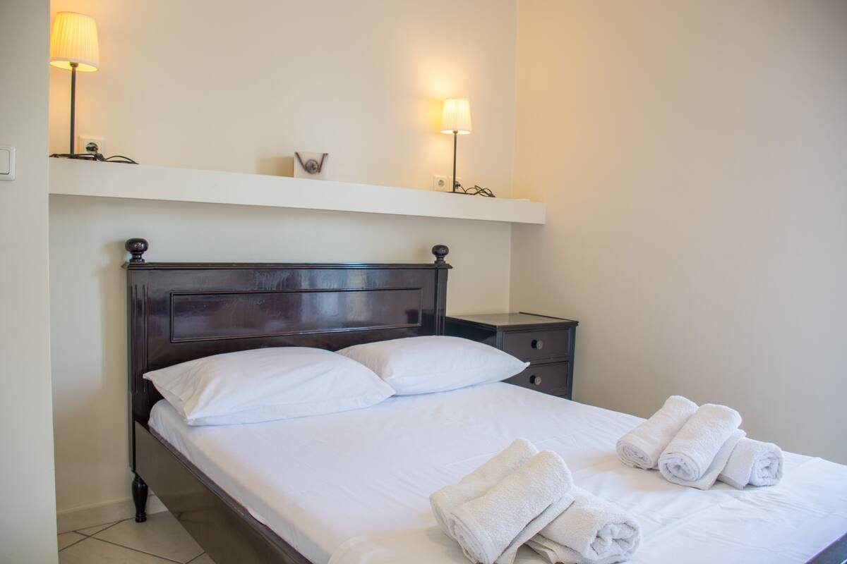 A neatly made bed with white linens and rolled towels, a dark wooden headboard, and two bedside lamps on a white shelf above.
