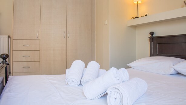 Quarto de hotel com cama, roupa de cama branca, guarda-roupa de madeira e uma luminária fixada na parede.