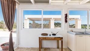 A kitchen with a white countertop, a wooden table, and a view of the landscape through large windows.