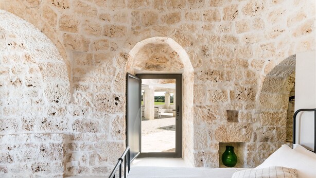 A stone-walled room with a bed, a green vase, and a view of an outdoor area.