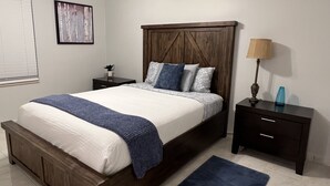 A bedroom with a wooden bed frame, a white mattress, and a blue throw blanket. There is a nightstand with a lamp and a blue vase. -  Spacious 1-bedroom just mins to  WEF  (Wellington)