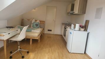 A compact, well-lit room with a bed, a desk with a chair, a kitchenette, and a door.