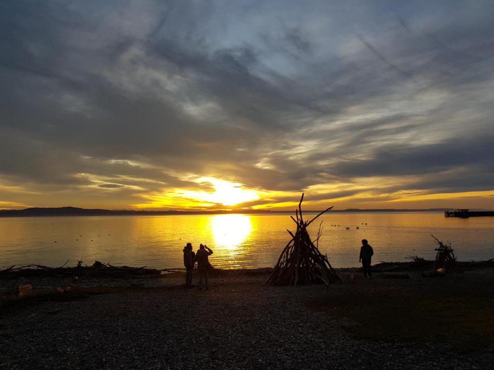 A sunset over a calm body of water with a group of people and a teepee-like structure on the shore.