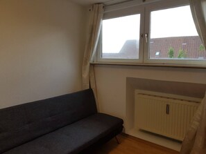 Ett rum med en mörkgrå soffa, ett fönster med gardiner och ett element.