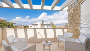 Uma área externa coberta com cadeiras brancas e uma pequena mesa, uma parede de pedra e vista para casas ao fundo.