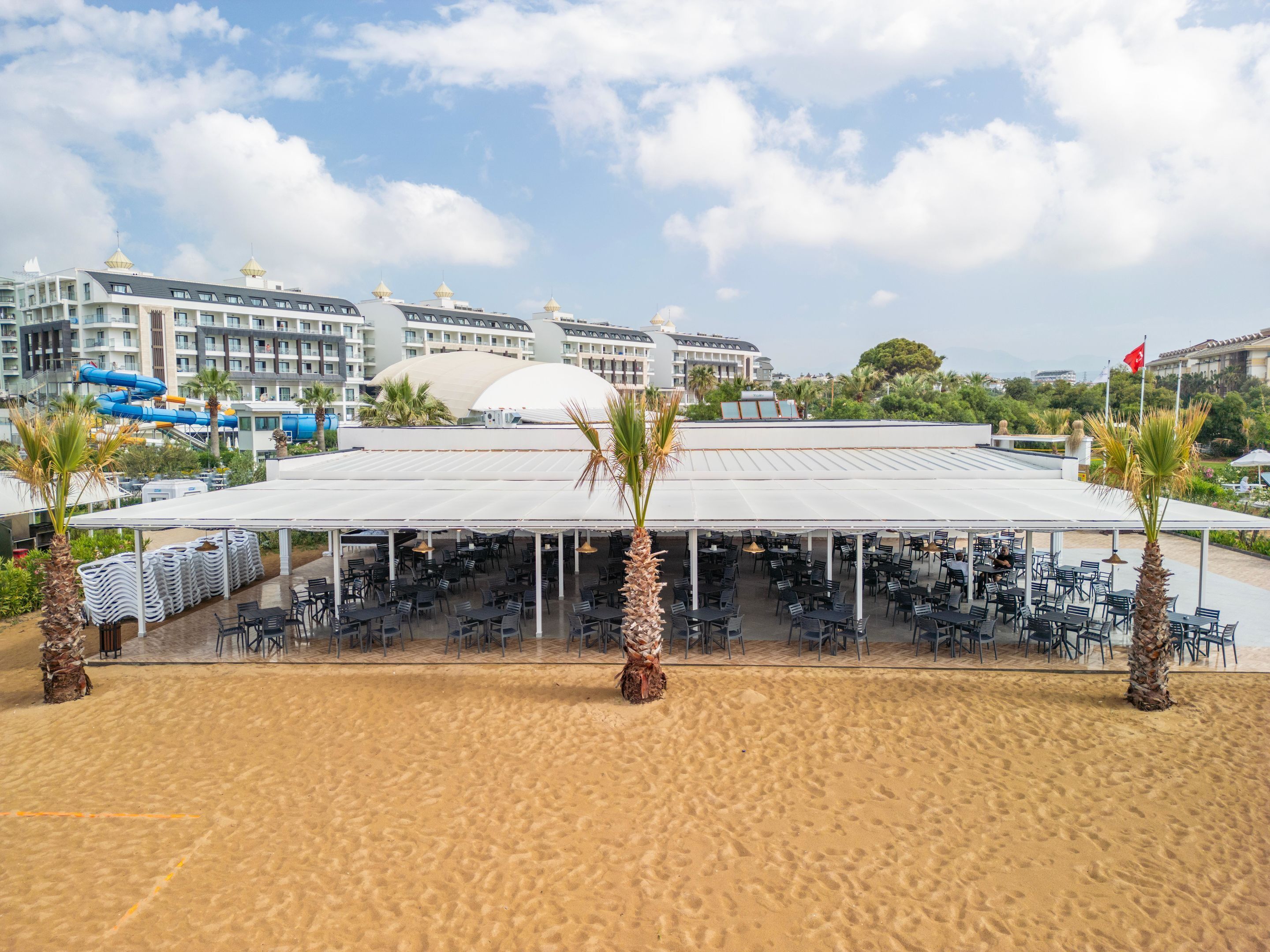 A beachside outdoor dining area with tables and chairs, palm trees, and a view of hotel buildings in the background.