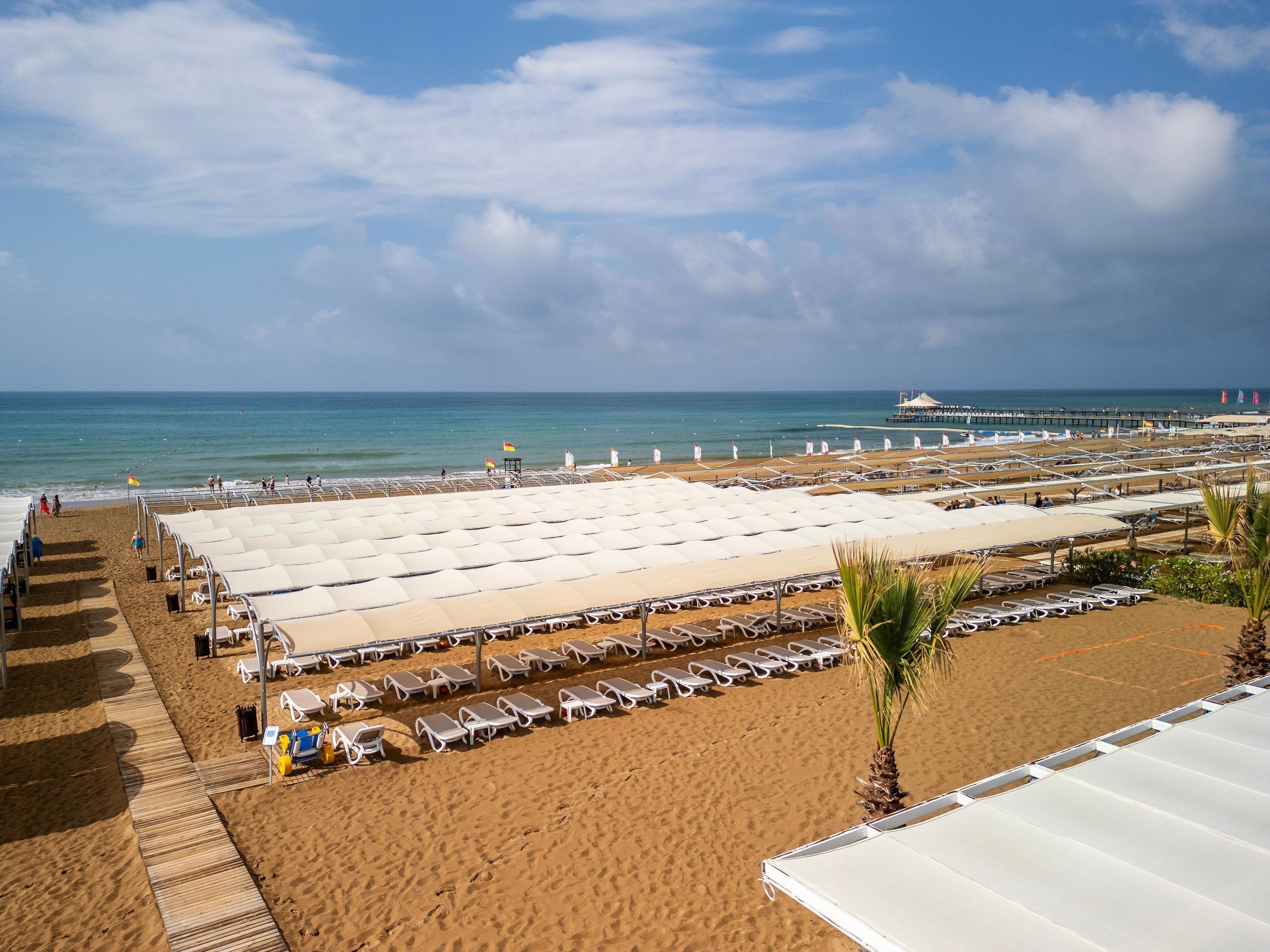 A beach with rows of sun loungers and umbrellas, a pier in the background, and a clear sky.
