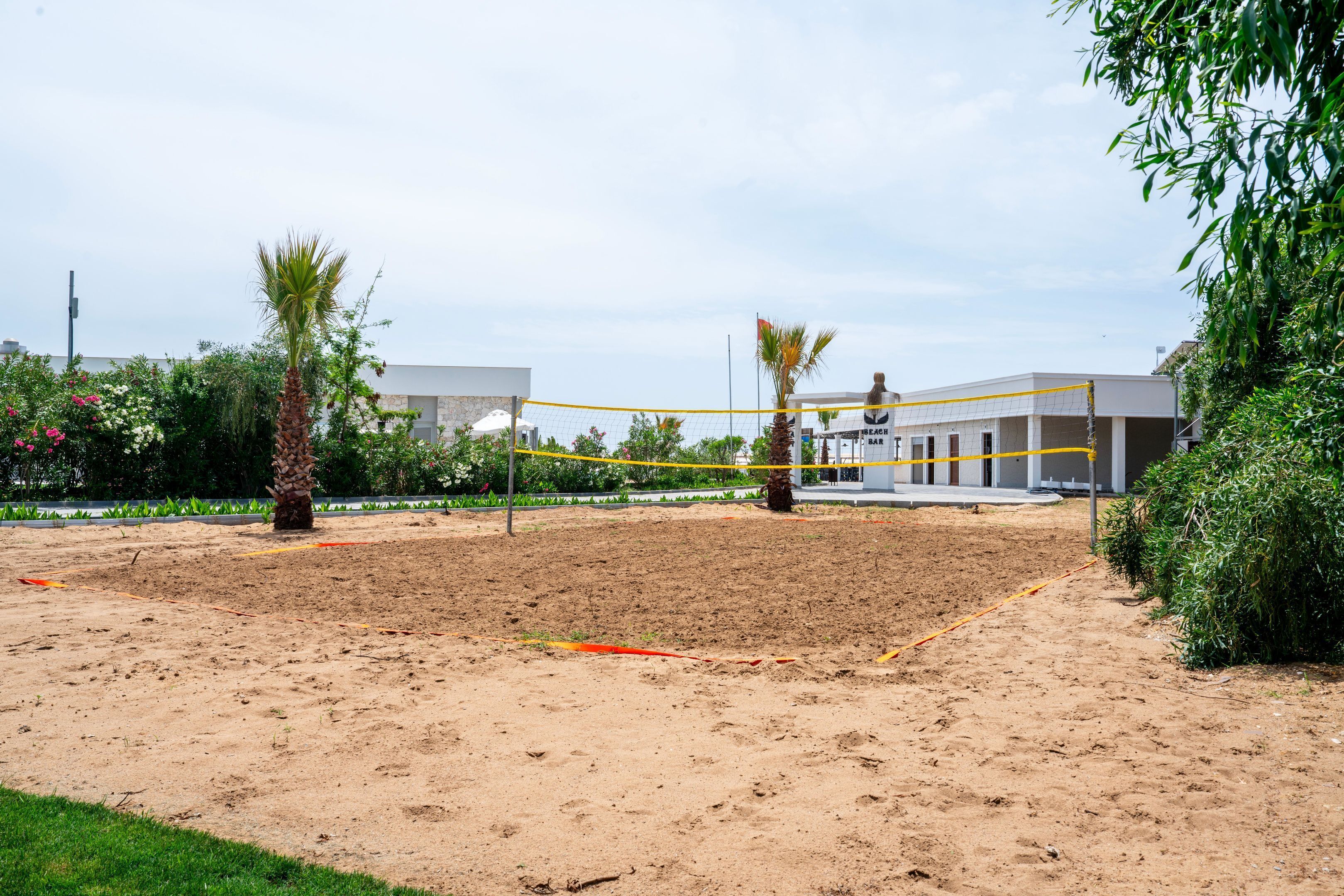 Uma quadra de vôlei de areia com rede, rodeada por vegetação e um edifício ao fundo.