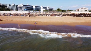 Uma praia com espreguiçadeiras e guarda-sóis, uma orla arenosa e edifícios ao fundo.