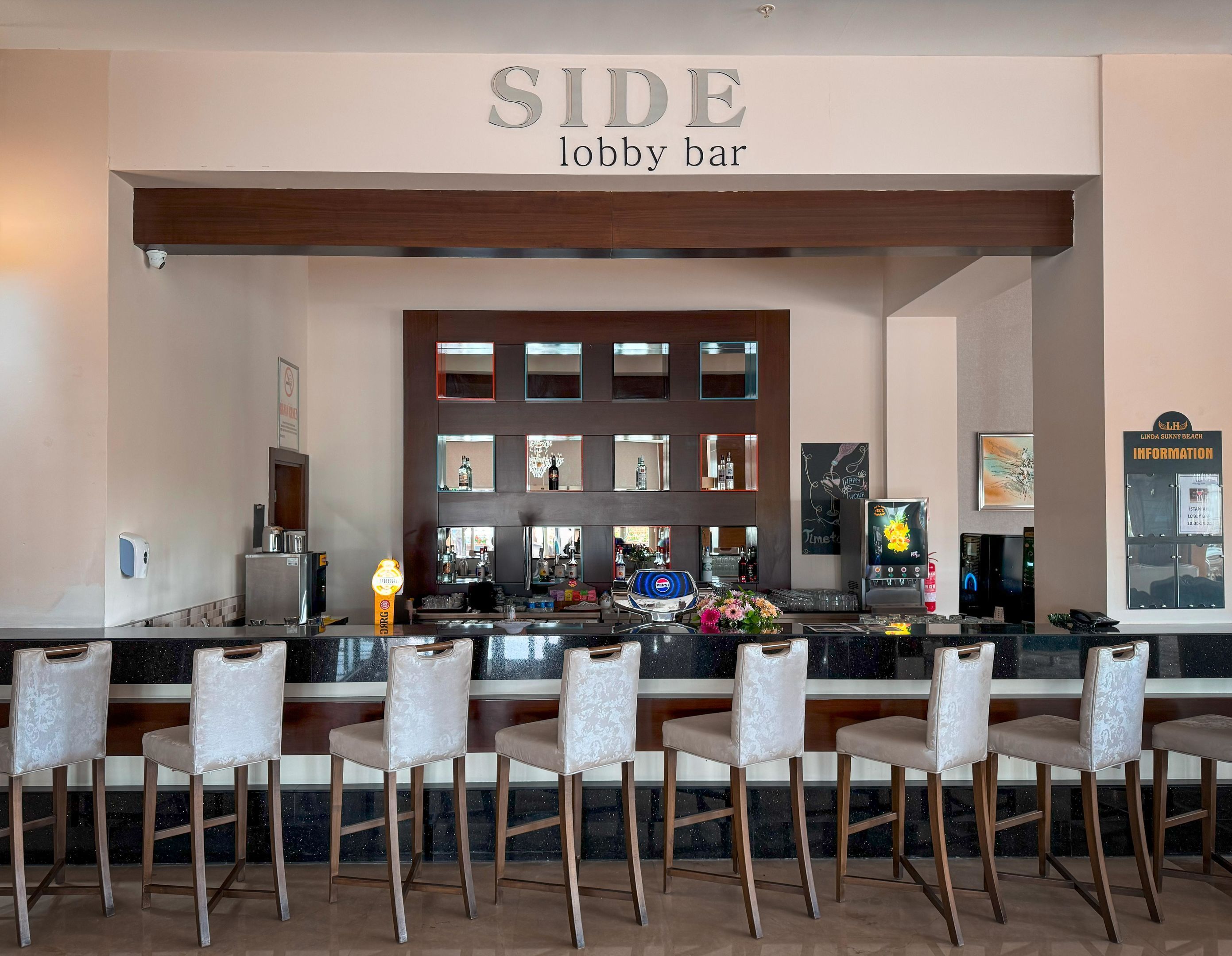 A lobby bar area with a sign reading "SIDE lobby bar," bar stools, a counter with a cash register, and a display of bottled beverages.