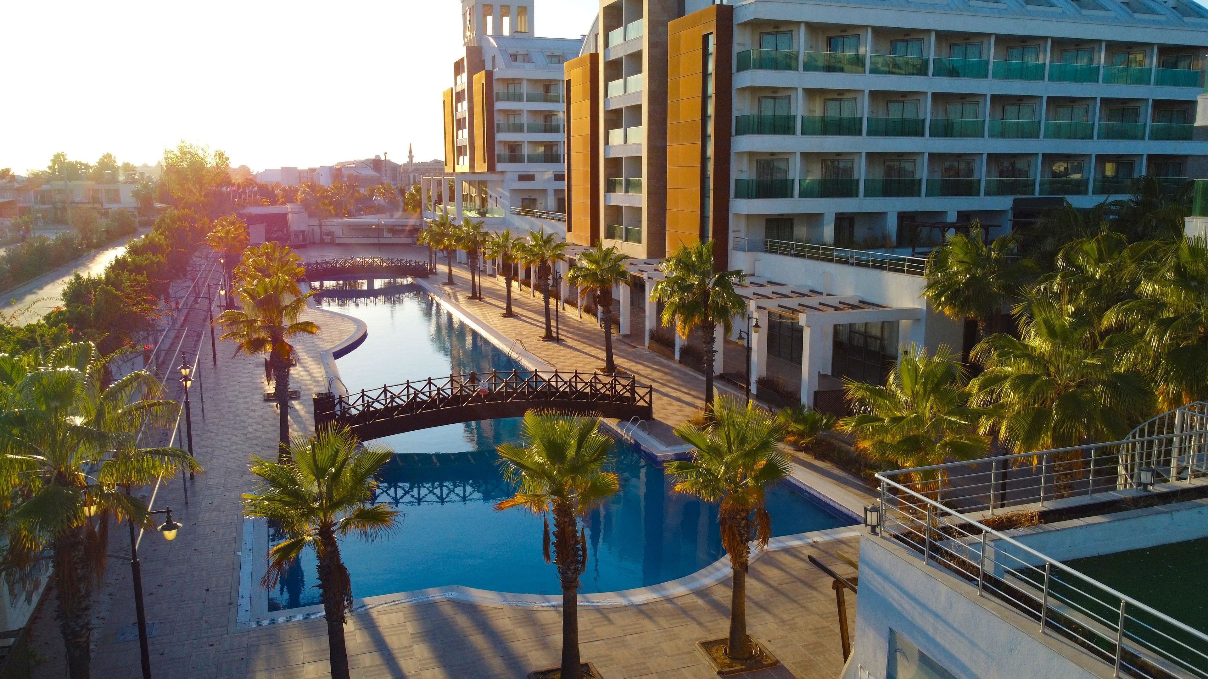 A modern hotel with a central swimming pool, surrounded by palm trees and a bridge.