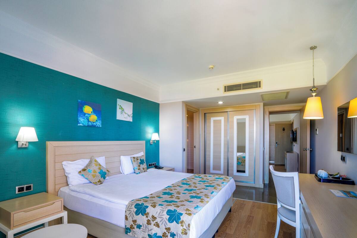 Camera d'albergo con un letto, comodini, una scrivania e una sedia. Si apre una porta verso il bagno e un corridoio.