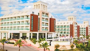 Um hotel com vários andares e design moderno, com entrada destacada e fachada de vidro, além de área frontal ajardinada com palmeiras.