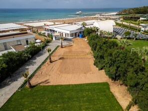 An aerial view of a beachside hotel with a sandy volleyball court, a parking area, and a view of the ocean.