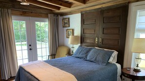 A bedroom with a large bed, wooden ceiling beams, a wooden door, and a view of greenery outside. - Remodeled French Country Style Cottage Close to Hiking Trails and Hot Springs (Hot Springs)