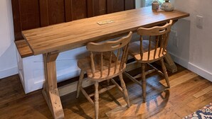 A wooden dining table with a bench, wooden chairs, a clock on the wall, and a window with a view of greenery. - Remodeled French Country Style Cottage Close to Hiking Trails and Hot Springs (Hot Springs)