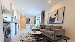 A modern apartment with a living area featuring a sofa, coffee table, and a dining table with chairs. There is a washing machine and a kitchen area visible. - MINT Apartments Greenside (Randburg)