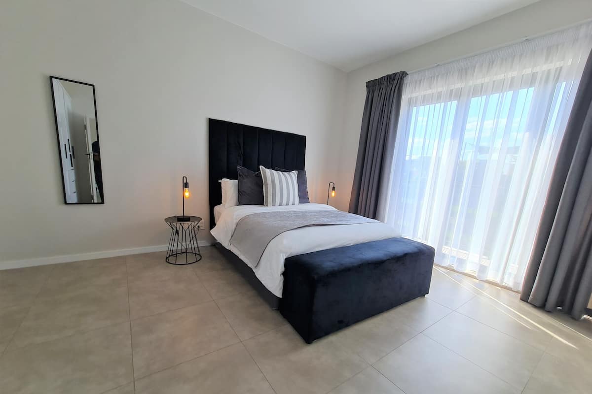 Een moderne slaapkamer met een groot bed, een rond nachtkastje, een zwart hoofdeinde en een groot raam met gordijnen.